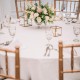Round table covered with a 90-inch white polyester tablecloth, adorned with a floral centerpiece for elegant dining.